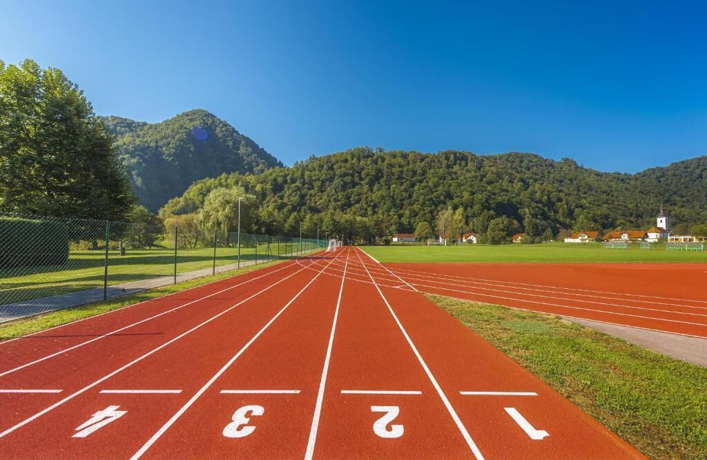 A running track with lane markings and a starting line, surrounded by a fence and trees, with a mountain in the background.