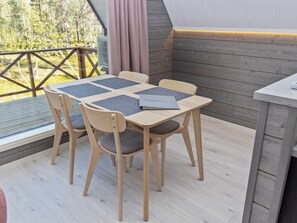 Una terraza de madera con una mesa y sillas, una ventana con vistas a árboles y una cortina.