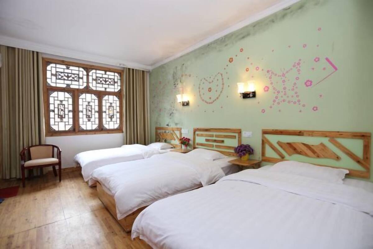 Habitación de hotel con tres camas, una silla de madera, un ventanal con detalles decorativos e iluminación en la pared.