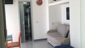 A modern living room with a sofa, a blue ottoman, a dining table with a chair, and a glass door leading to a bathroom.