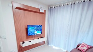 Een moderne woonkamer met een flatscreen televisie bevestigd aan een houten wandpaneel, een eethoek met een tafel gedekt voor twee personen, en een bank met decoratieve kussens.