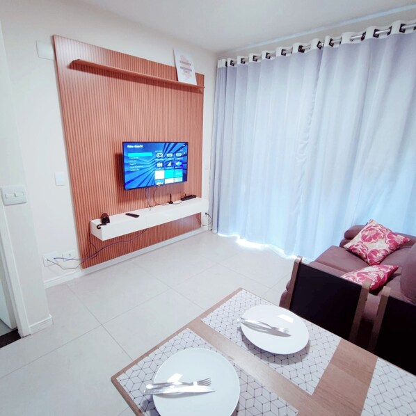 Ein modernes Wohnzimmer mit einem Flachbildfernseher an einer Wandpaneele aus Holz, einem Essbereich mit Tisch für zwei Personen und einer Couch mit Zierkissen.
