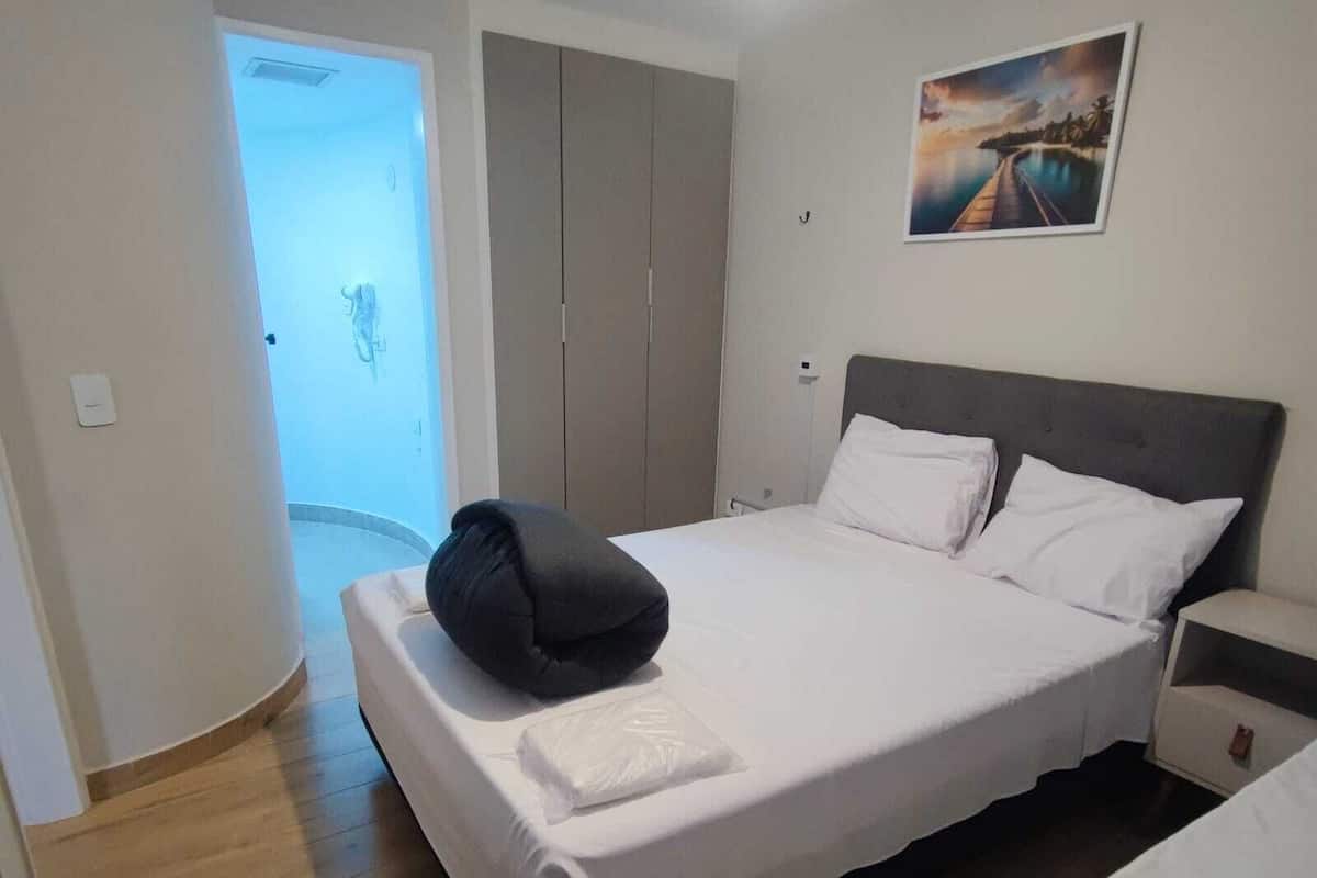 Een opgemaakt bed met witte lakens en een zwart kussen, een nachtkastje met een lamp, en een schilderij aan de muur.