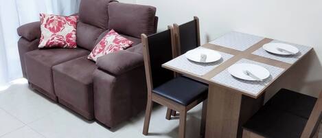 Een woonkamer met een bruine bank, twee bijpassende stoelen, een klein houten tafeltje met een tafelkleed met ruitpatroon, en een wit bord met een vork erop.