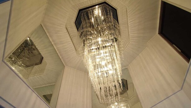 Een modern interieur met een grote, versierde kroonluchter die aan het plafond hangt, omringd door geometrische wandpanelen en een groot raam.