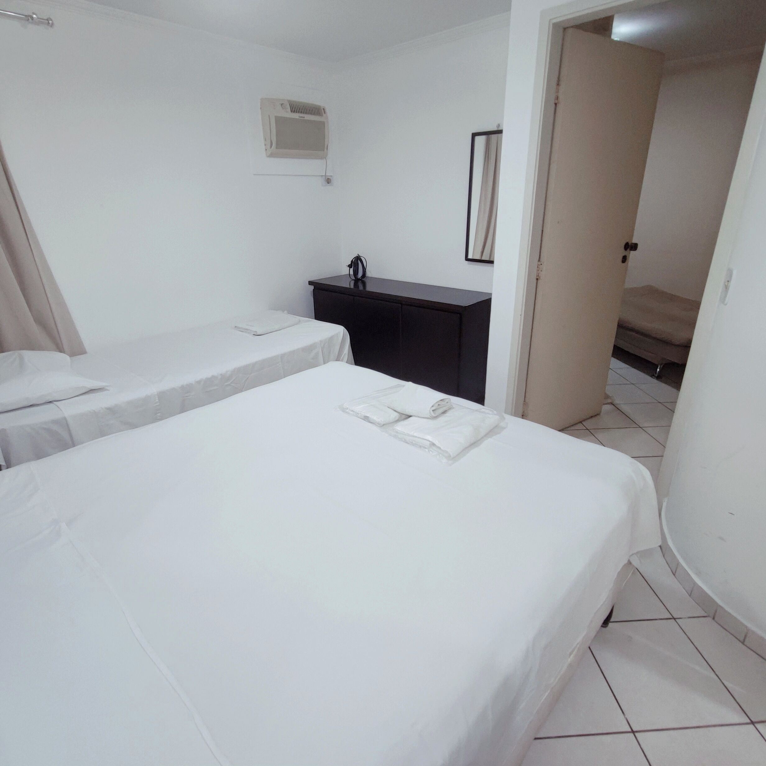 Habitación de hotel con dos camas, un aparador de color oscuro, un equipo de aire acondicionado pequeño en la pared y una puerta que da a otra habitación.