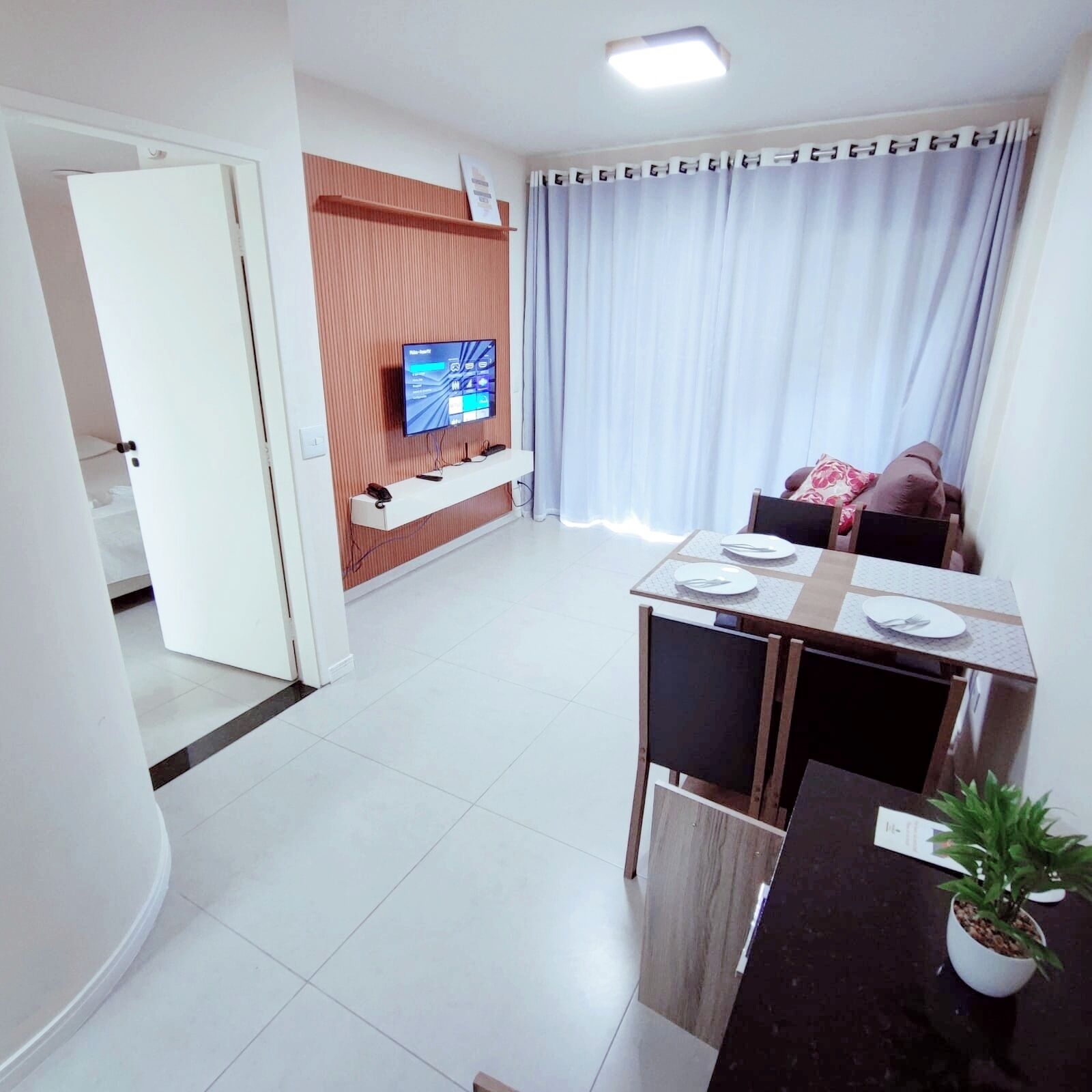 A modern living room with a flat-screen TV mounted on a wooden panel, a dining area with a table set for two, and a potted plant on a dark countertop.