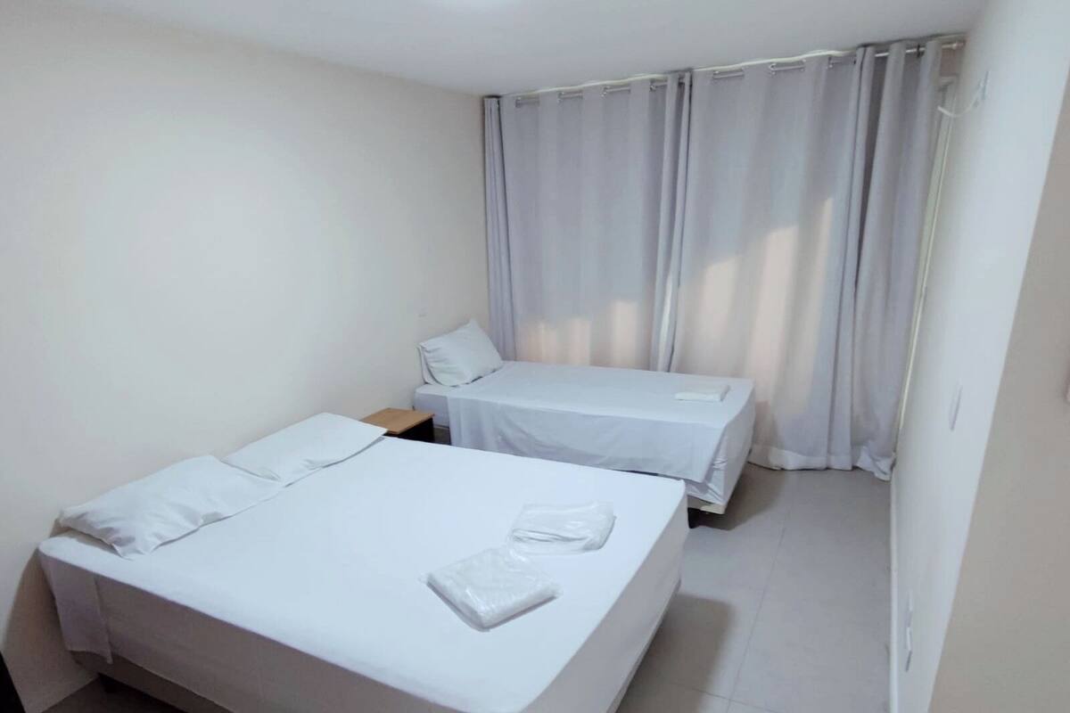 Een hotelkamer met twee bedden, witte lakens, een klein nachtkastje en een raam met gordijnen.