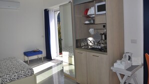 A compact bedroom with a bed, a small table, a kitchenette with a microwave, and a sliding door leading to a balcony.