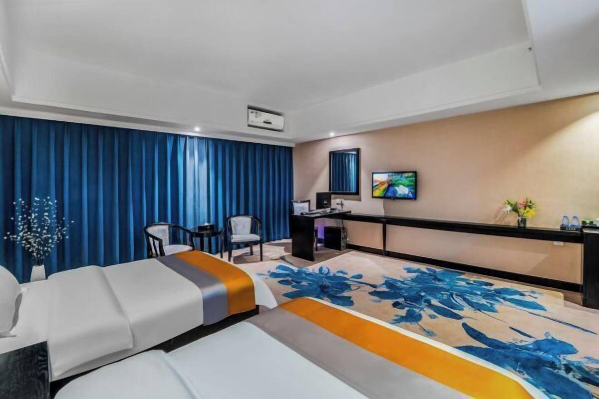 A hotel room with two beds, a desk, a chair, a television, and a large window with blue curtains.
