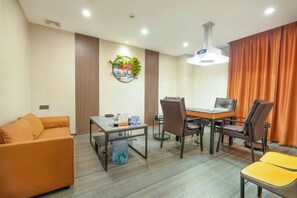 Habitación de hotel con sofá, una mesa de comedor con sillas, una mesita con lámpara y un ventanal con cortinas.