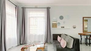 Una sala de estar moderna con un sofá gris oscuro, una mesa de centro de vidrio y una mesita auxiliar de madera. Hay cortinas en las ventanas y un letrero de bienvenida en la pared.