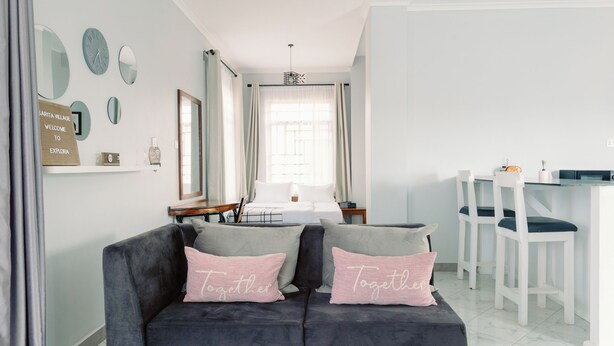 A modern living room with a dark grey sofa, two pink pillows, and a white dining area with bar stools. - Cozy studio suite with a full kitchen and sitting area (Arusha)
