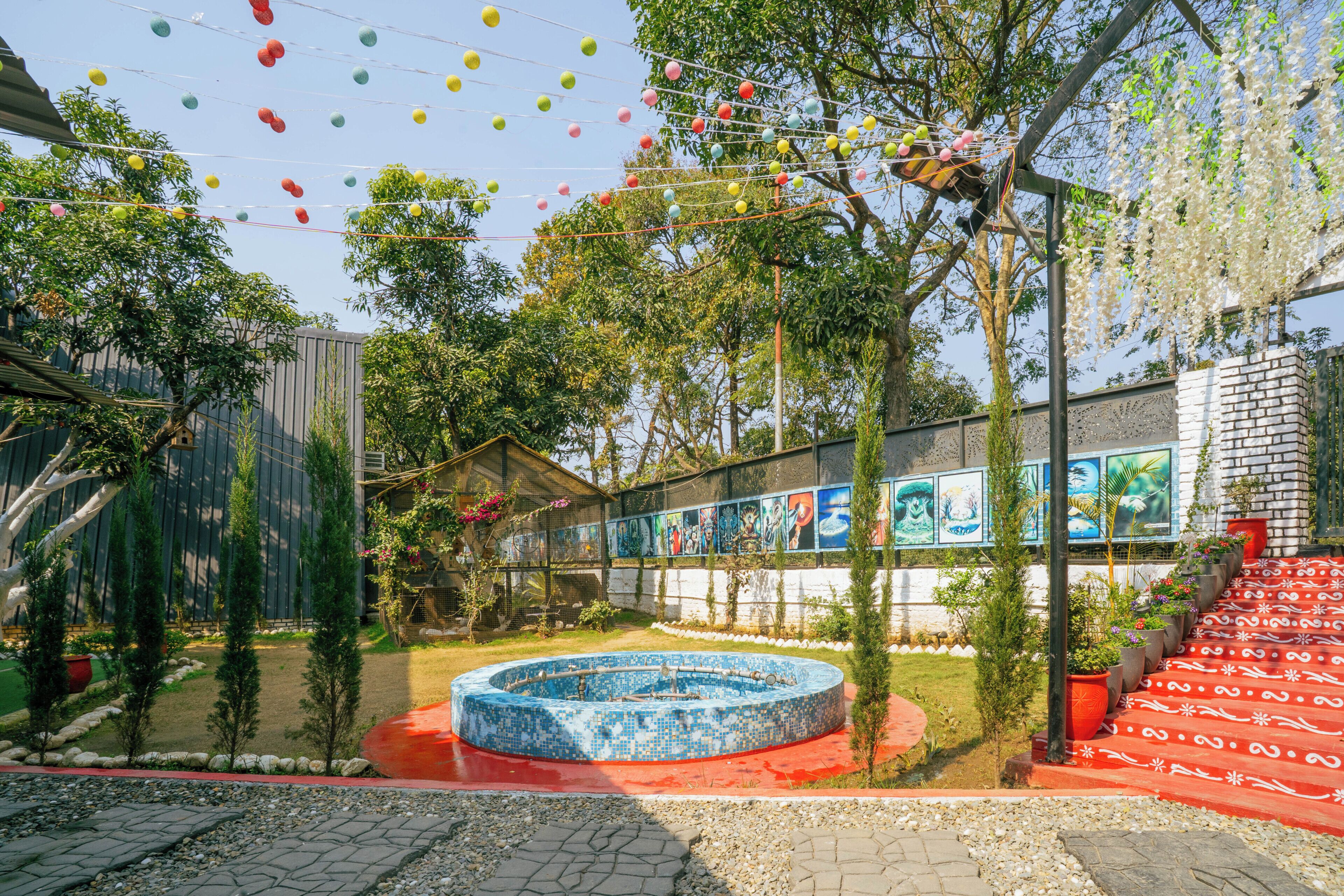 Une cour avec une piscine ornée d’une mosaïque circulaire, un escalier aux marches rouges et un bâtiment décoré d’une fresque murale.