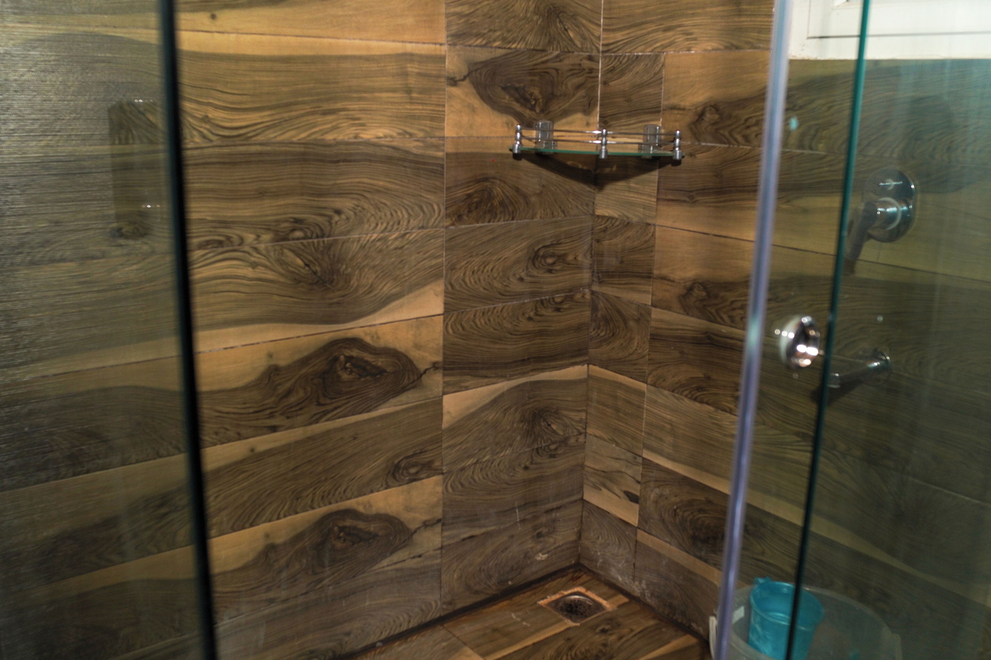 A shower with a glass door, wooden panel walls, and a small shelf with a glass container.