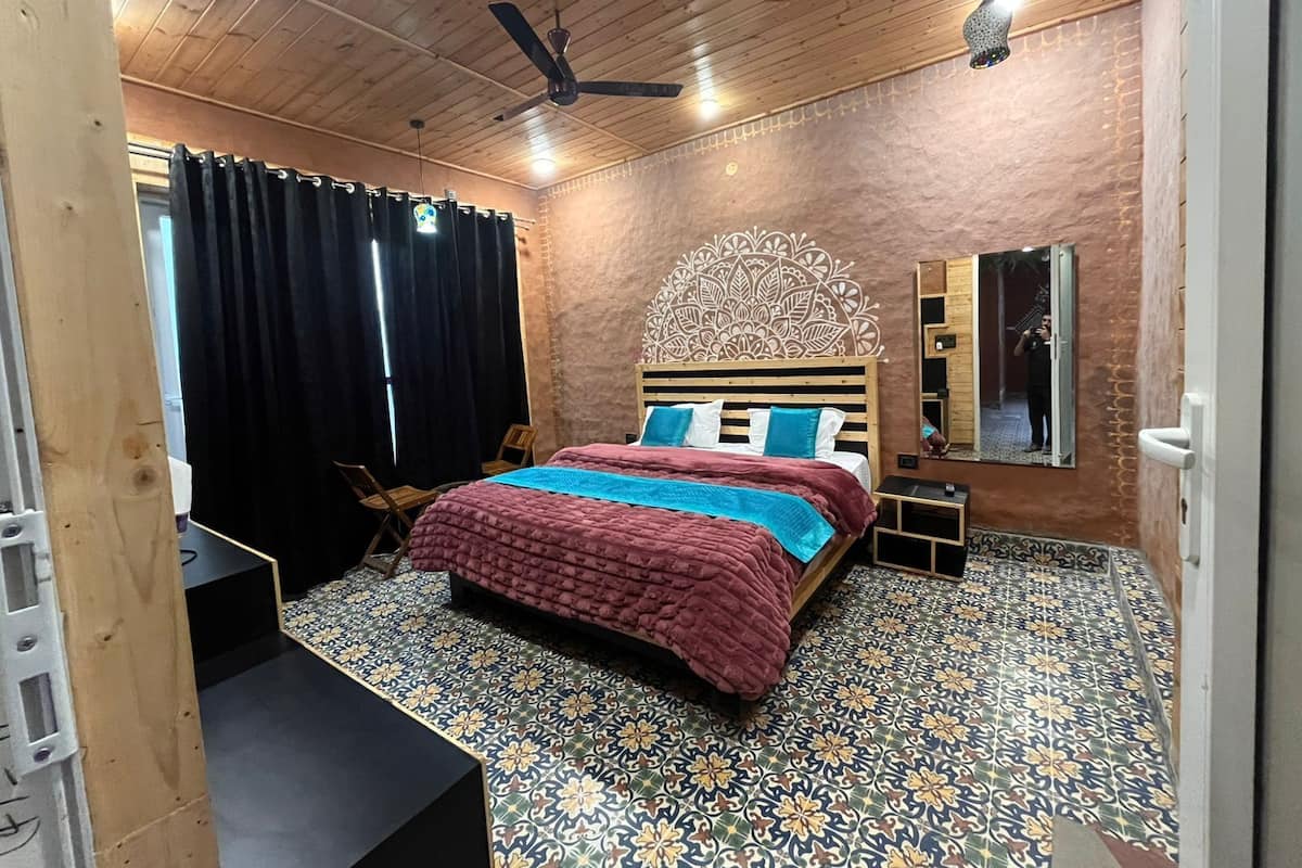 Ein Schlafzimmer mit Holzbalkendecke, einem Bett mit gemustertem Überwurf, einem dekorativen Kopfteil, einem Nachttisch, einem Spiegel und einem Fenster mit Vorhängen.