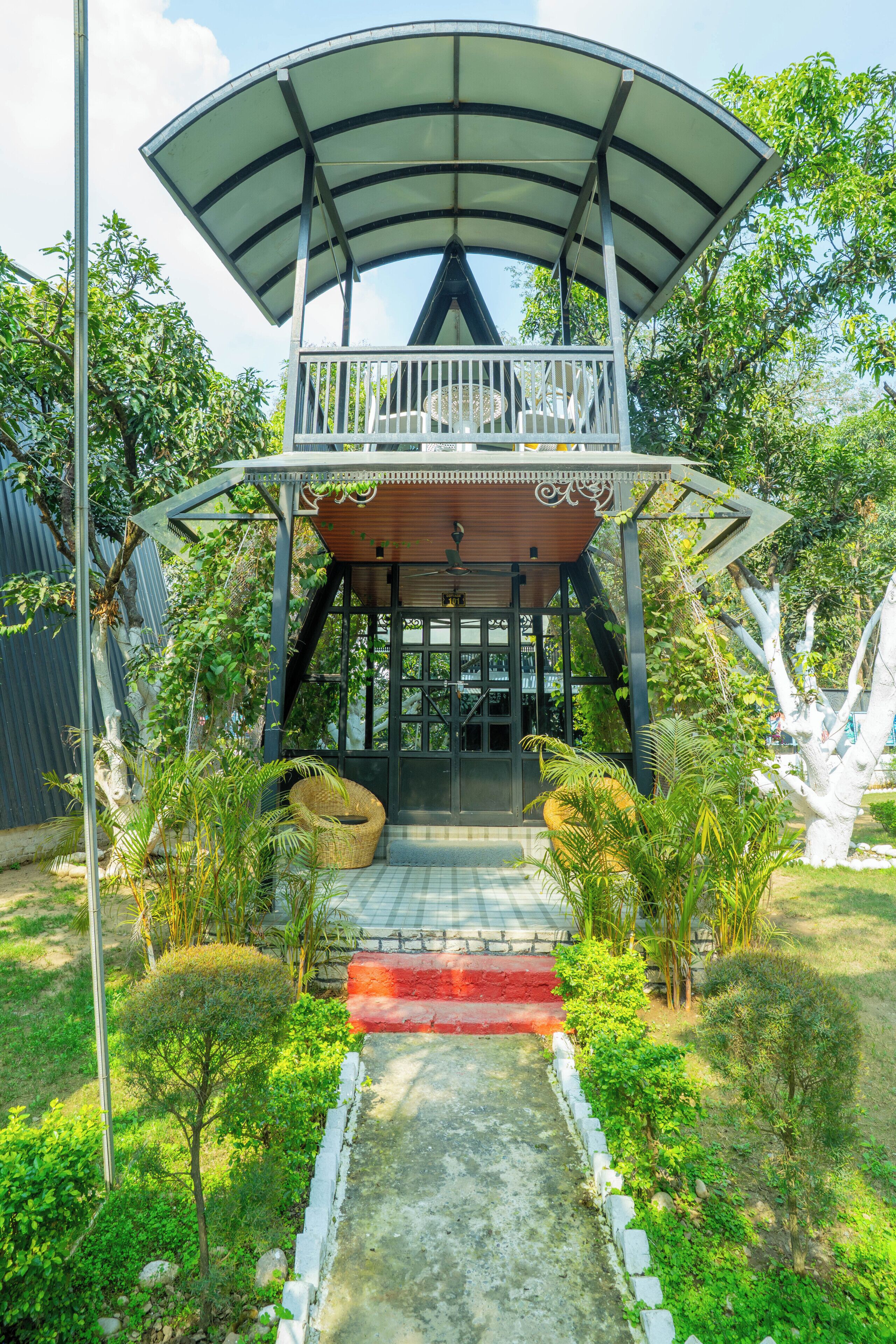 A two-story structure with a covered balcony, surrounded by greenery and a concrete pathway.