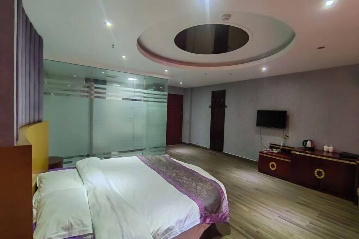 A hotel room with a bed, a TV, a wooden dresser, and a glass partition wall.