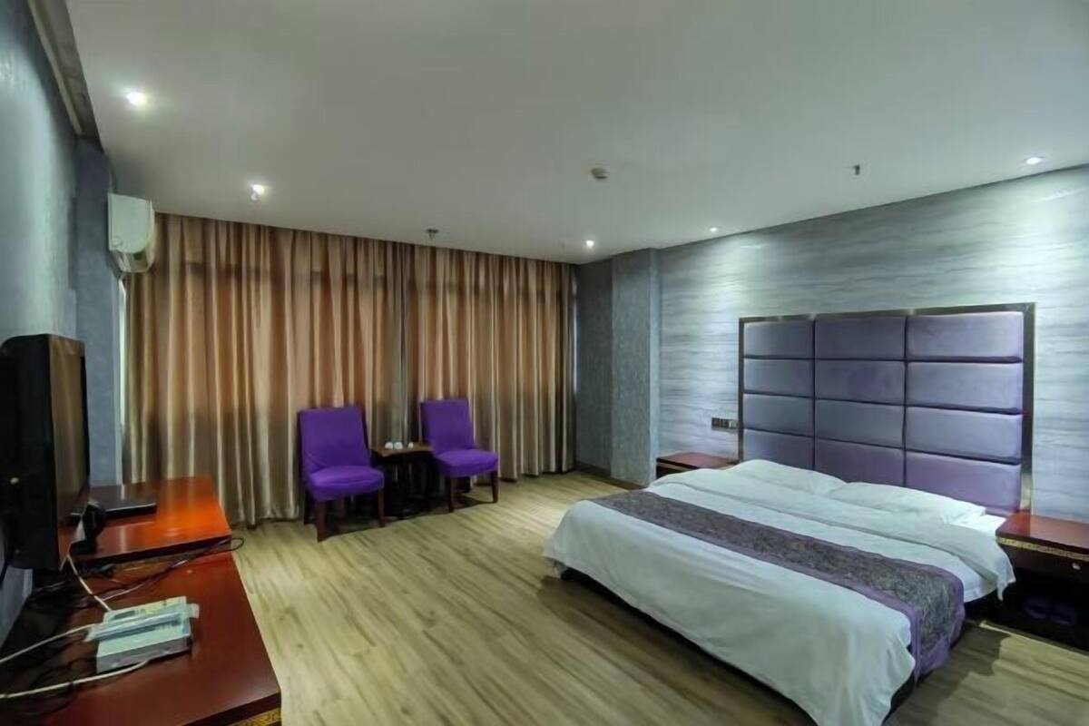 A hotel room with a bed, two purple chairs, a TV, and a wooden floor.