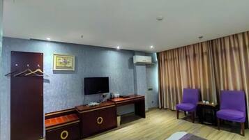 Habitación de hotel con un escritorio de madera, un televisor, dos sillas moradas, un armario y una cortina.