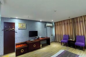 Habitación de hotel con un escritorio de madera, un televisor, dos sillas moradas, un armario y una cortina.