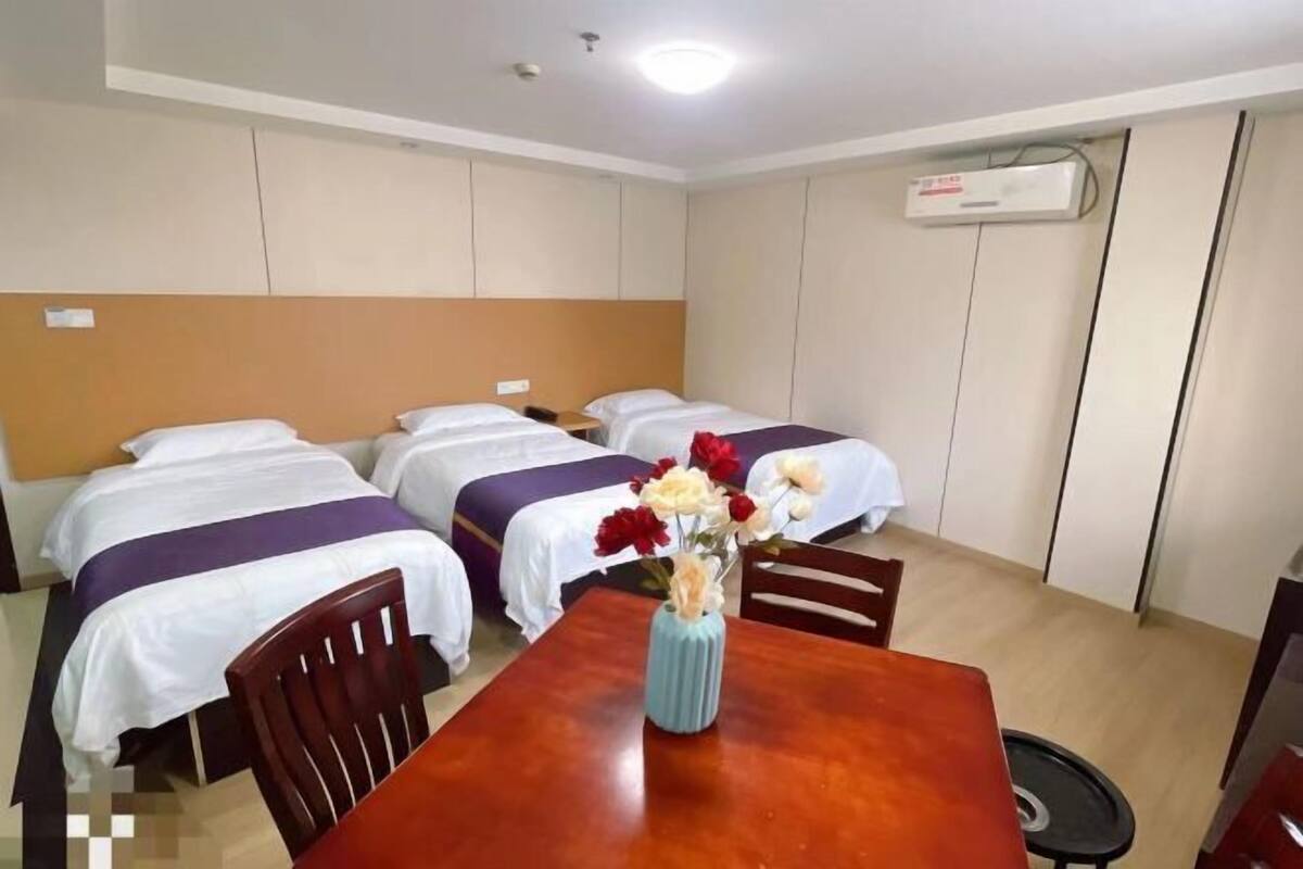 Habitación de hotel con tres camas, una mesa de madera con un florero y un equipo de aire acondicionado en la pared.