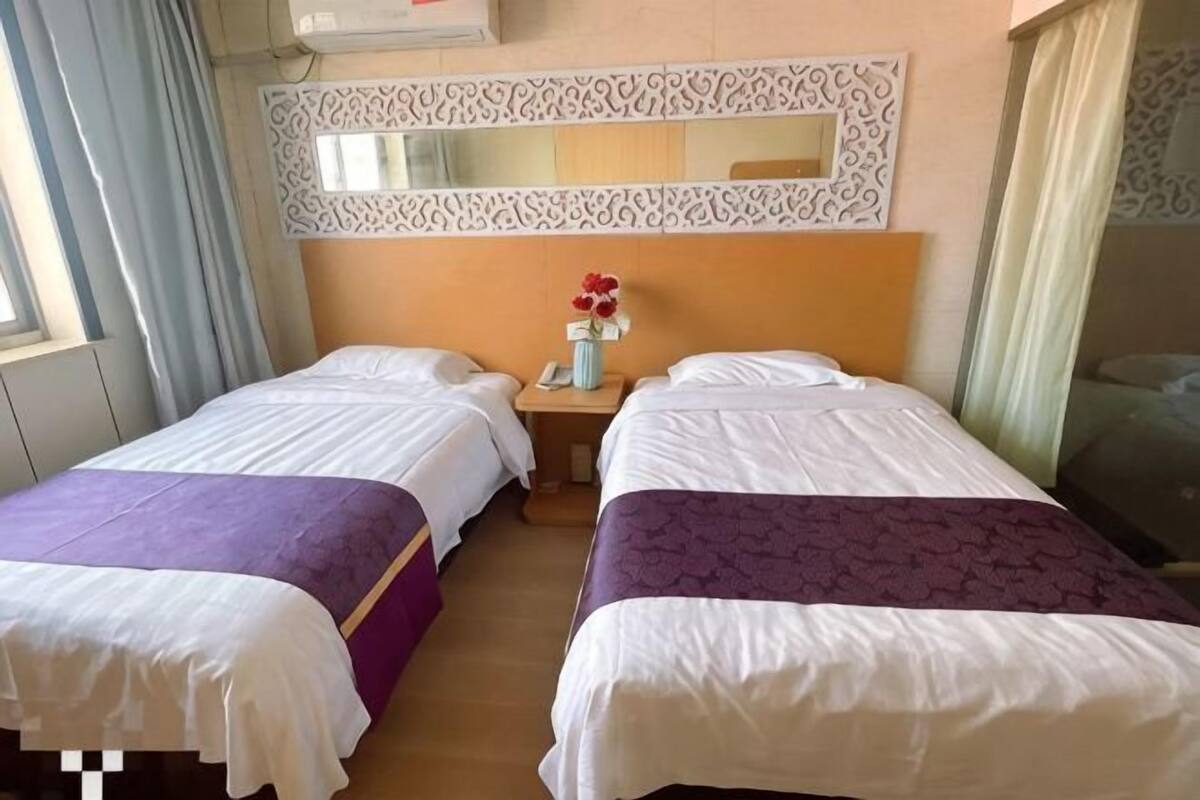 Habitación de hotel con dos camas, una mesita de noche con un jarrón de flores, un espejo sobre las camas y una ventana con cortinas.