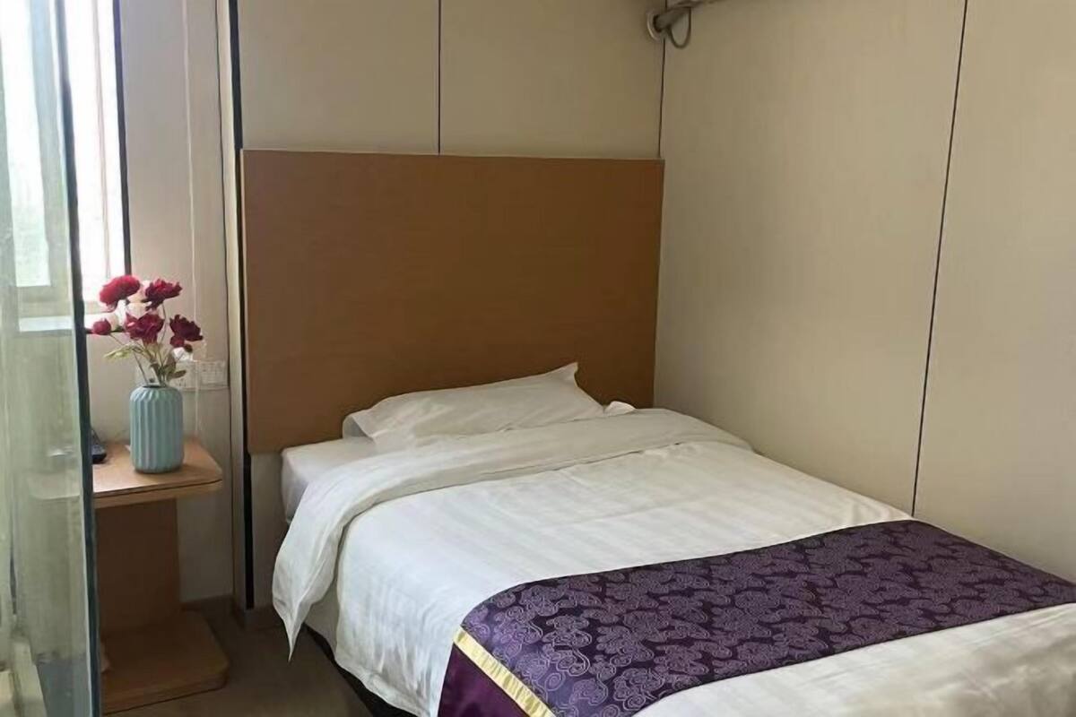 Una cama individual con cabecera, una mesita de noche con un florero y una ventana con cortinas.