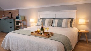 A neatly made bed with a tray of champagne, glasses, and snacks, a bedside table with a lamp, a microwave, and a small shelf with a toaster.