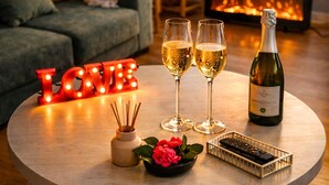 A cozy living room with a fireplace, a round table with two glasses of champagne, a bottle of wine, and a decorative 'LOVE' sign.