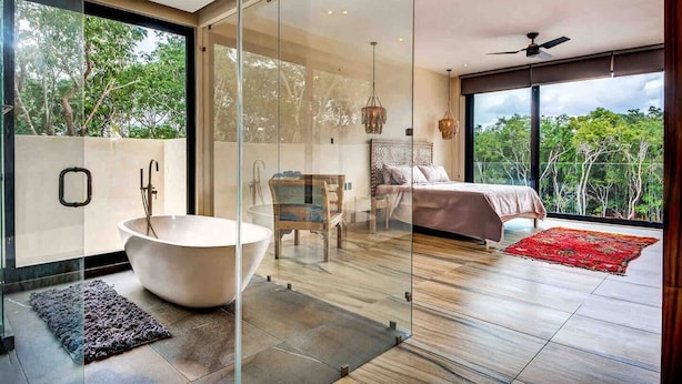 A modern bedroom with a large glass door, a bathtub, a bed, and a balcony with a view of trees. - Tulum Dream Villa: Designer Luxury Near The Beach (Tulum)