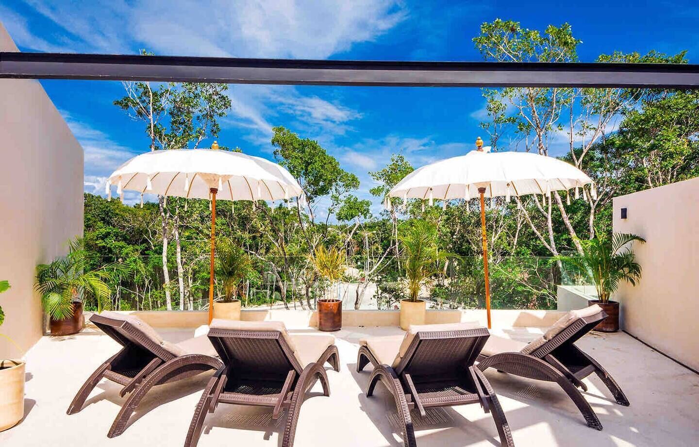 A rooftop terrace with two lounge chairs and two umbrellas, surrounded by lush greenery and trees.