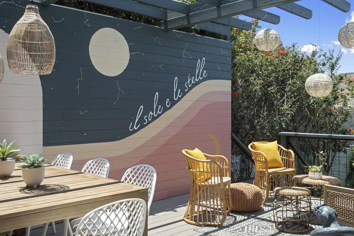 An outdoor seating area with a wooden table and wicker chairs, a mural with text, and hanging paper lanterns.
