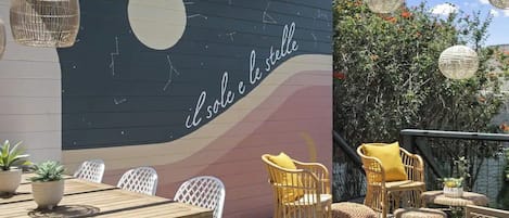 An outdoor seating area with a wooden table and wicker chairs, a mural with text, and hanging paper lanterns.