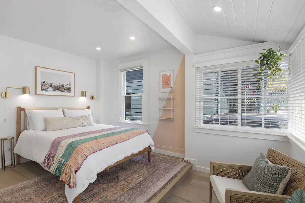 Un dormitorio con cama, mesitas de noche, una ventana con persianas y una planta colgante.