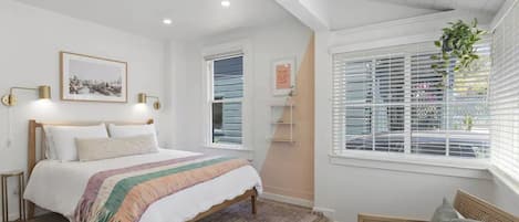 Un dormitorio con cama, mesitas de noche, una ventana con persianas y una planta colgante.