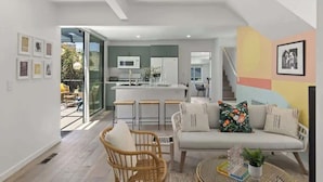 A modern living room with a sofa, a wicker chair, and a glass coffee table. There is a kitchen area with a microwave and a refrigerator in the background. - 5BR (Sleeps 12!)  Downtown Little Italy Home w/ Outdoor Space (San Diego)