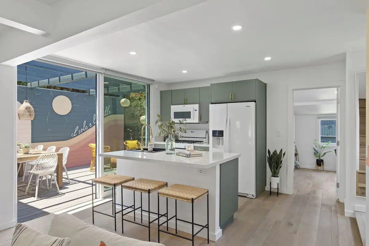 Una cocina moderna con una isla central, muebles blancos y una zona de comedor con sillas de mimbre.