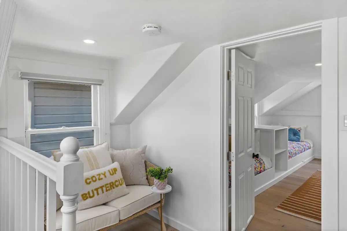 Una habitación con escalera blanca, un banco empotrado con cojines, una mesita con una planta y una cama litera con ropa de cama colorida.