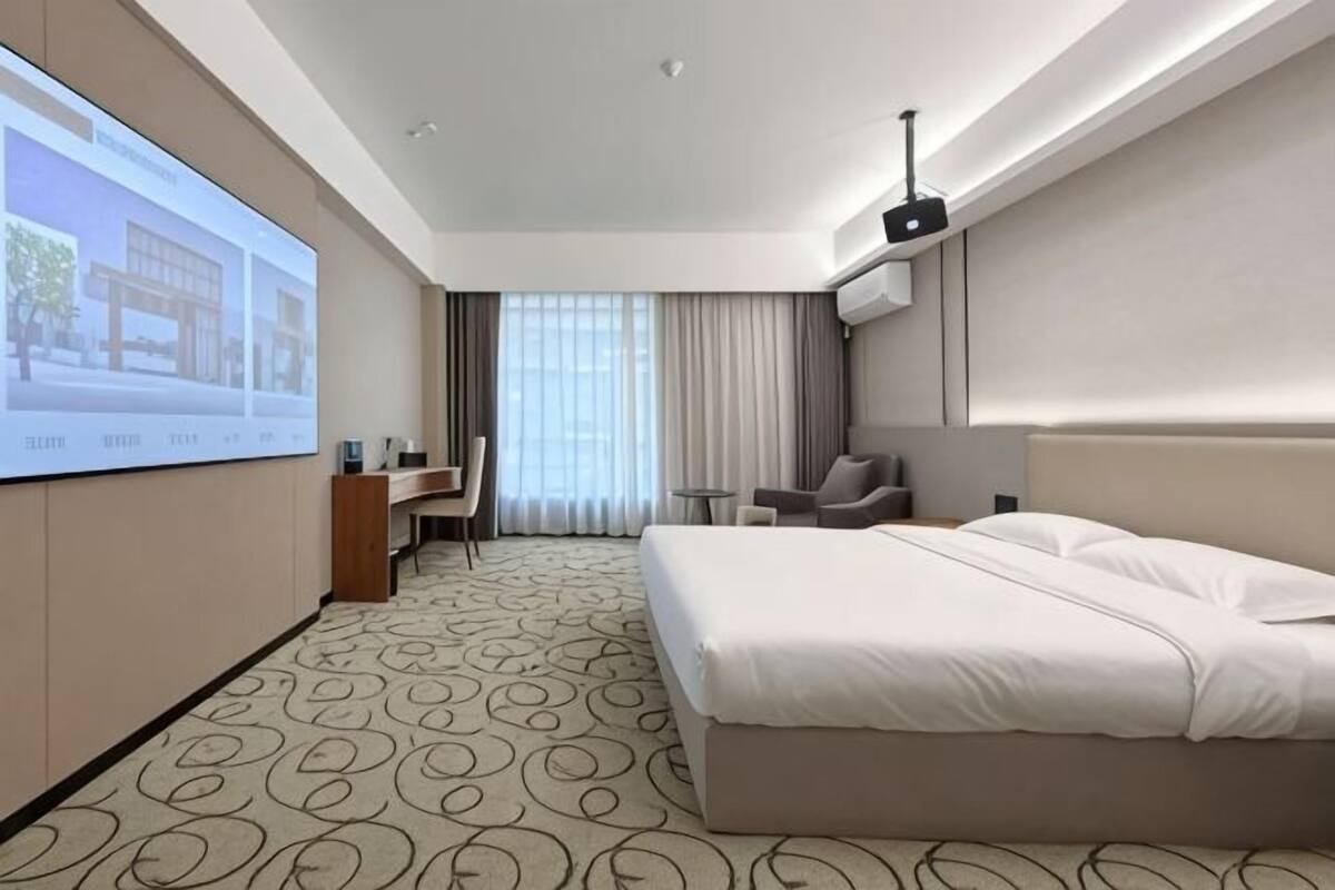Une chambre d’hôtel avec un grand lit, un bureau avec une chaise, une petite table, un écran de télévision et une grande fenêtre avec des rideaux.