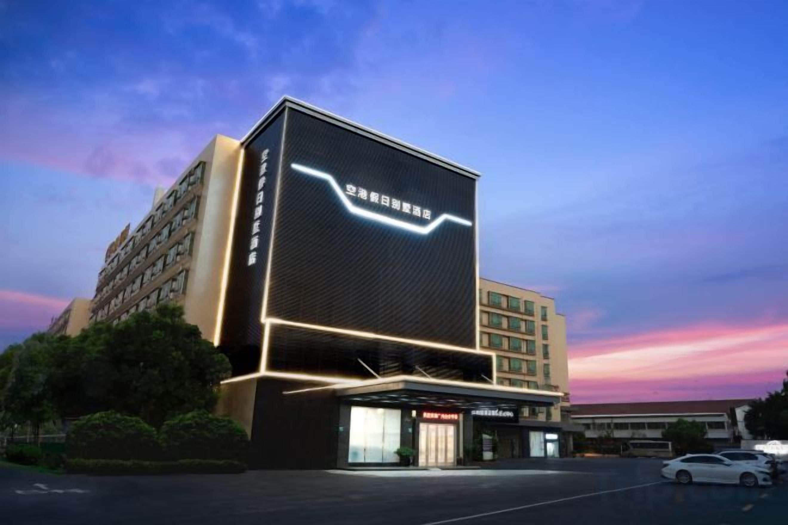 A modern hotel with a prominent digital display on the facade, a parking lot, and a clear sky.