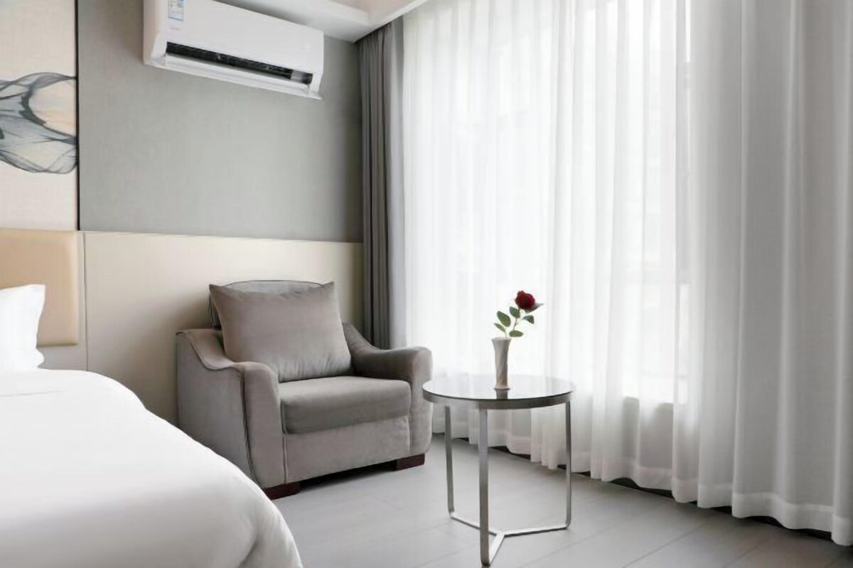 Een hotelkamer met een bed, een grijze fauteuil, een tafeltje met een vaas, en dunne gordijnen.