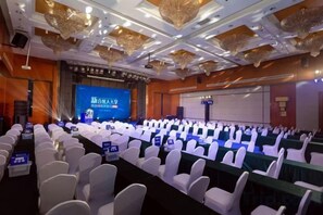 Une salle de conférence avec des rangées de chaises blanches, une scène dotée d’un écran de présentation et un plafond agrémenté d’éléments décoratifs.