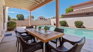 Un patio exterior con mesa de madera y sillas de mimbre, piscina y vistas a casas vecinas.