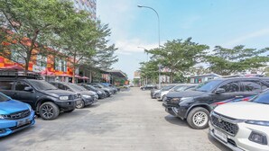 Un parking où de nombreuses voitures sont garées en rangées, des arbres bordent la rue et des bâtiments se dressent à l’arrière-plan.