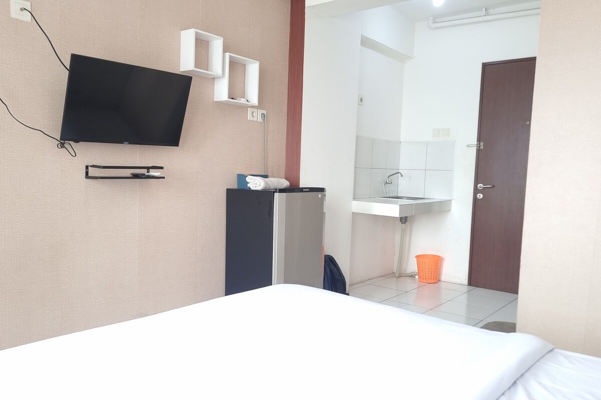 A hotel room with a bed, a TV mounted on the wall, a small desk with a chair, a mirror, a sink, and a door.