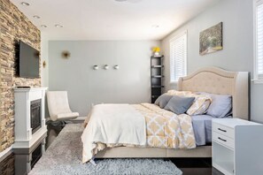 Un dormitorio con cama, una silla, televisor, chimenea y ventana con persianas.