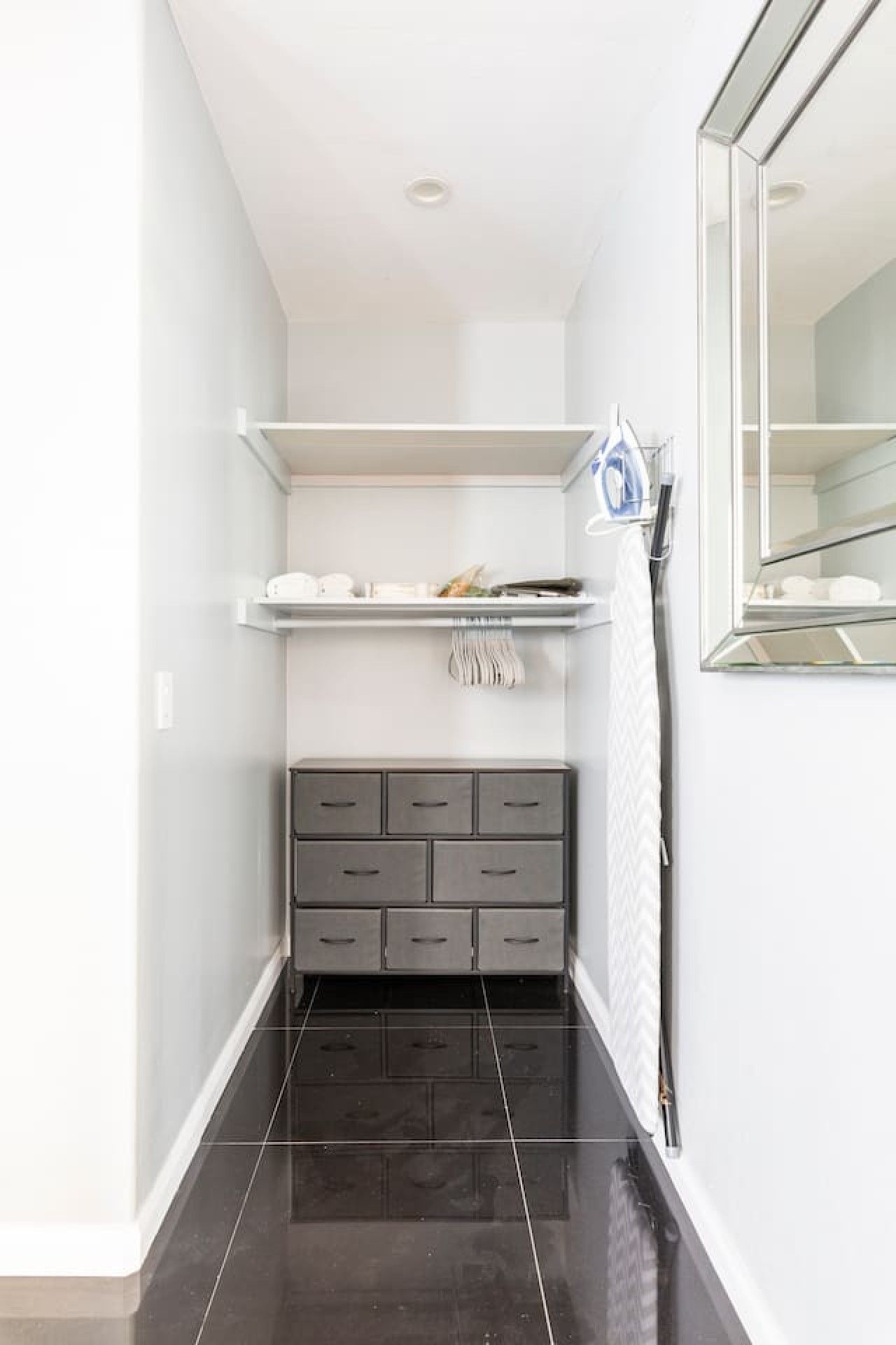 Un couloir étroit avec un sol carrelé foncé, une armoire grise avec des tiroirs, une étagère blanche sur laquelle sont posés divers objets, et un miroir sur le mur de droite.