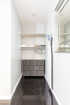 A narrow hallway with a dark tiled floor, a grey cabinet with drawers, a white shelf with items, and a mirror on the right wall.