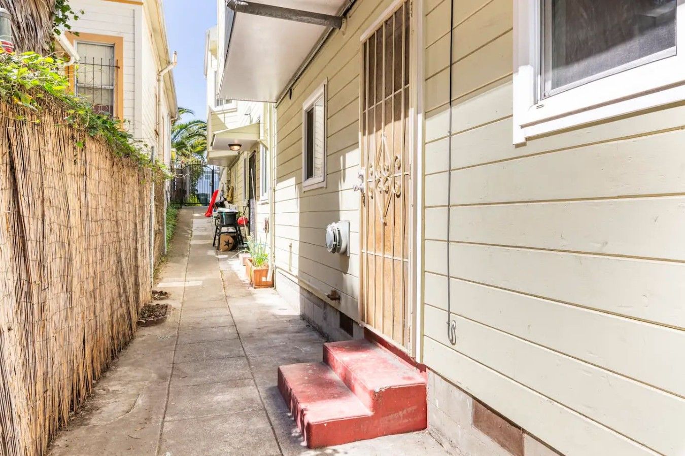 Une ruelle étroite, située entre deux bâtiments, avec une clôture en bois d’un côté et un bâtiment blanc de l’autre, devant lequel se trouve une marche rouge menant à une porte.